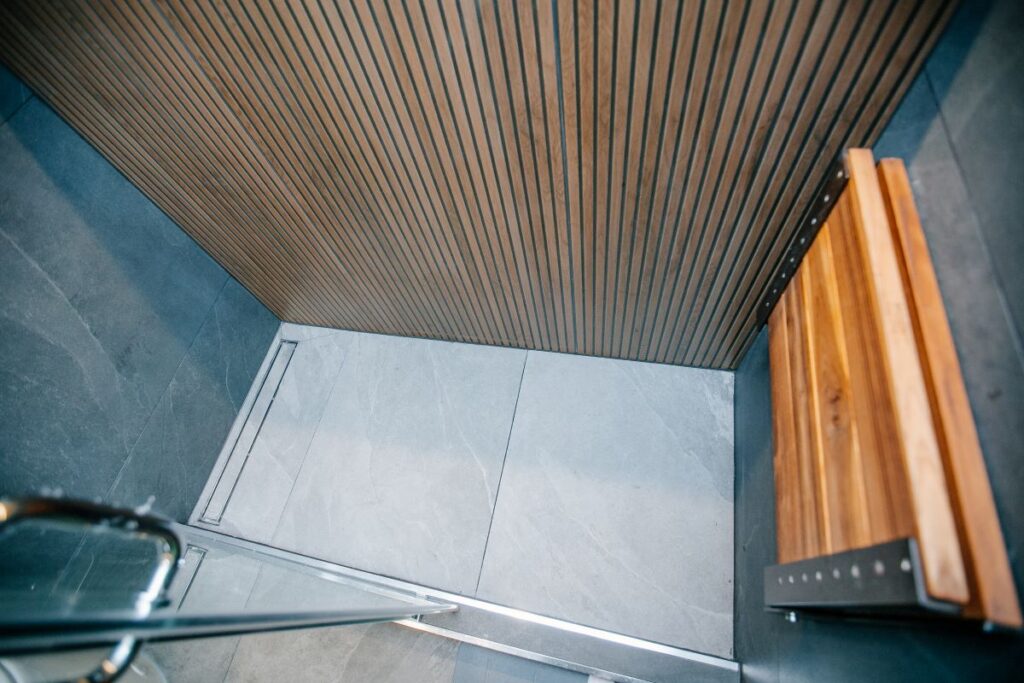 Top down view of a walk in shower with backlit oak slat wall, folding teak bench and a linear drain in grey porcelain