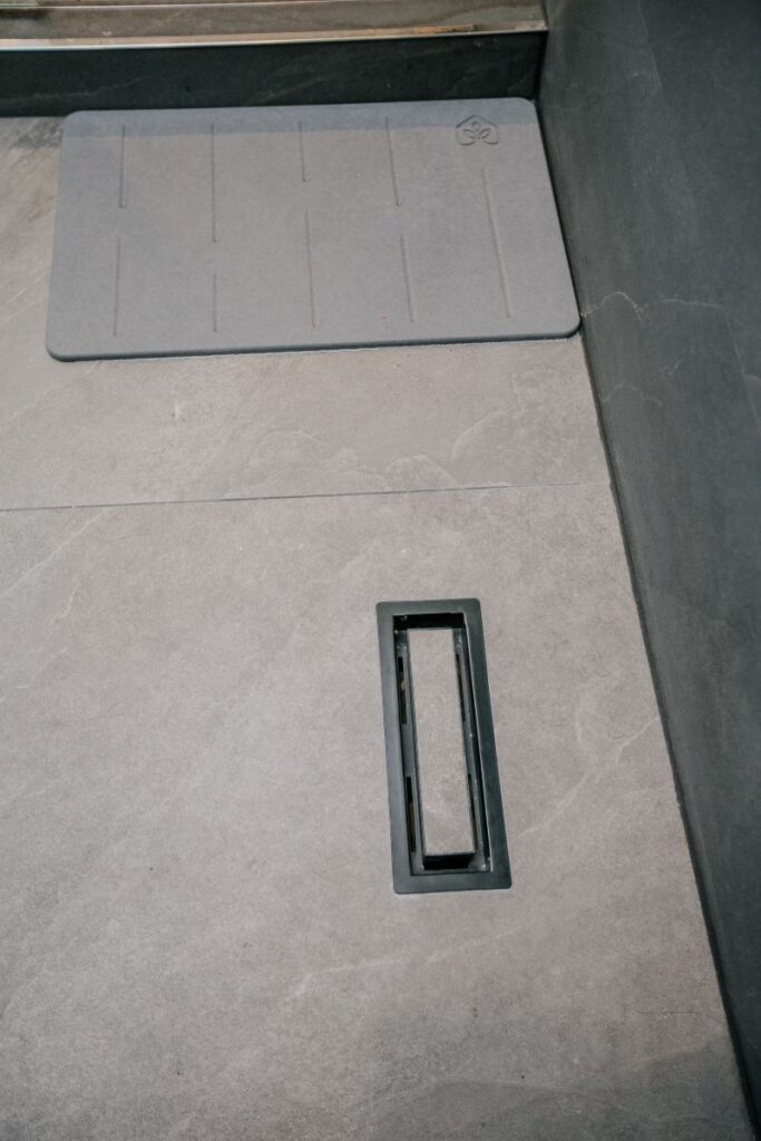 Walk in shower floor with a matte black square drain, grey stone look porcelain and an anti slip bath mat