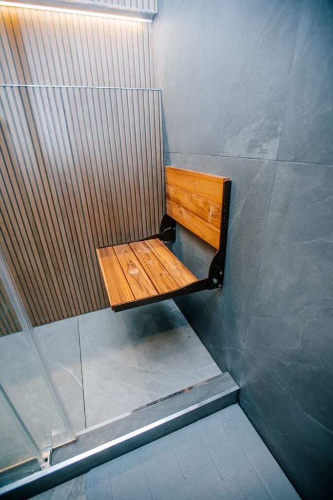 Wall mounted folding teak shower bench in a walk in shower with grey slate look porcelain and a backlit oak slat feature wall